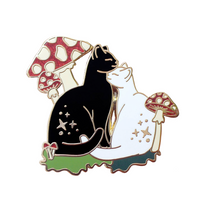 An enamel pin of a black cat and white cat cuddling amongst red and white mushrooms