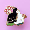 An enamel pin of a black cat and white cat cuddling amongst red and white mushrooms