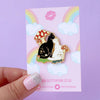 An enamel pin of a black cat and white cat cuddling amongst red and white mushrooms