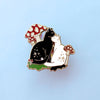 An enamel pin of a black cat and white cat cuddling amongst red and white mushrooms