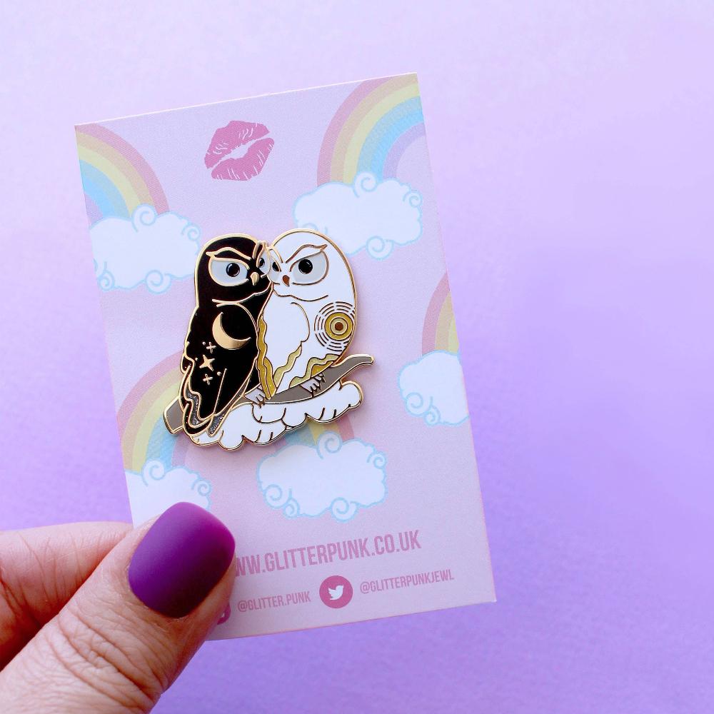 Enamel pin of a black owl representing night cuddling with a white owl representing day on packaging