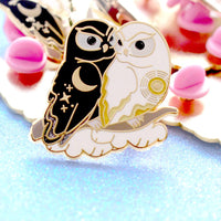 Closeup of enamel pin of a black owl representing night cuddling with a white owl representing day